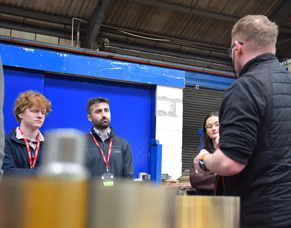 A group of three attentive employees wearing lanyards listen to a colleague speaking in an industrial workshop or manufacturing facility.
